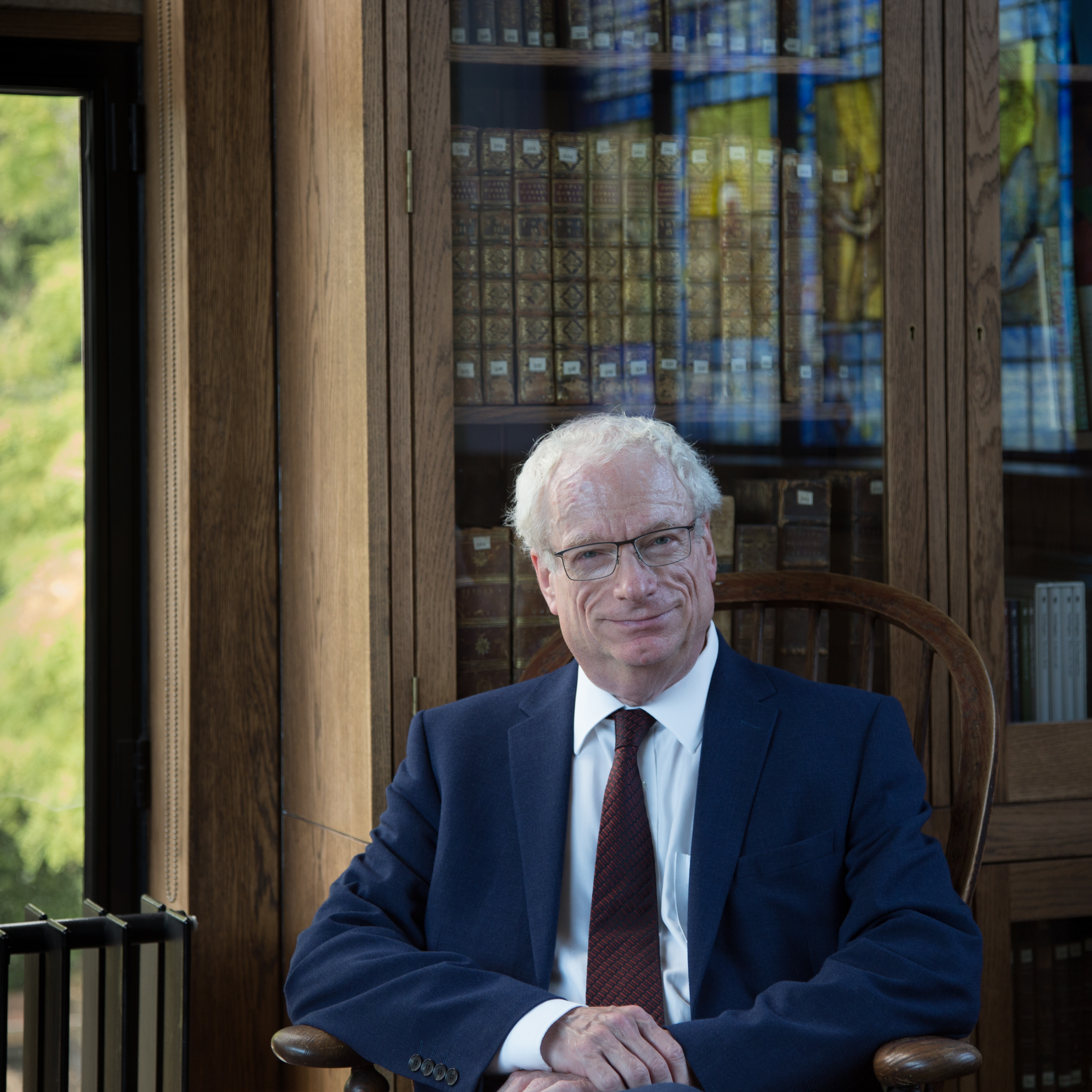 Photograph of Lord Chris Smith sitting in a chair in front of a bookcase. To the left of the photo is a window through which greenery can be seen .