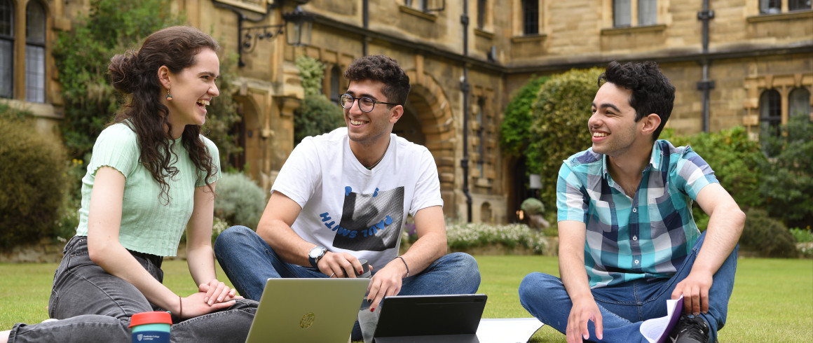 three students sat chatting on a lawn with their books and laptops