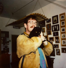 Michael Pritchard, dressed in a in brown suede jacket and blue shirt, holding a cat 