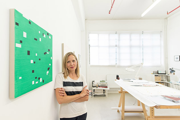 Artist Alison Turnbull leaningon a white wall, nextto one of her paintings